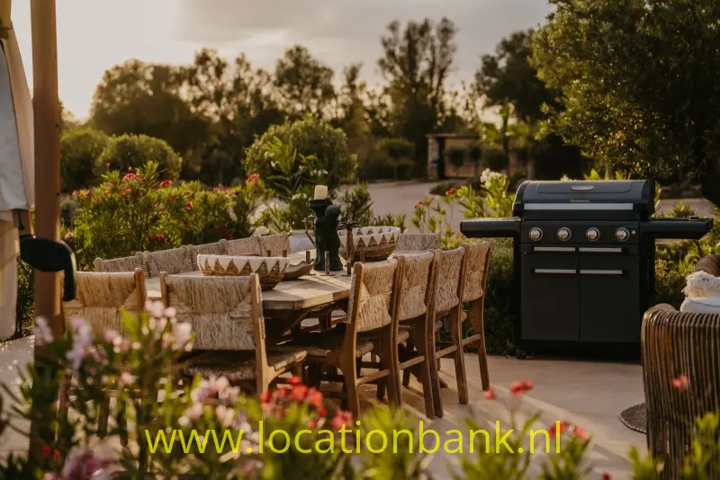 Een zwarte barbecue staat naast een lange houten eettafel met gevlochten stoelen, omringd door bloeiende planten.