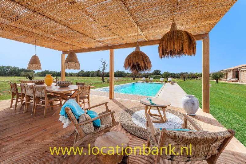 Overdekt terras met gevlochten hanglampen, houten eettafel en ligstoelen naast een rechthoekig zwembad.