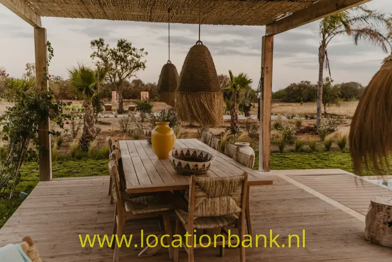 Een houten eettafel op een overdekt terras met gevlochten hanglampen en een groen landschap op de achtergrond.