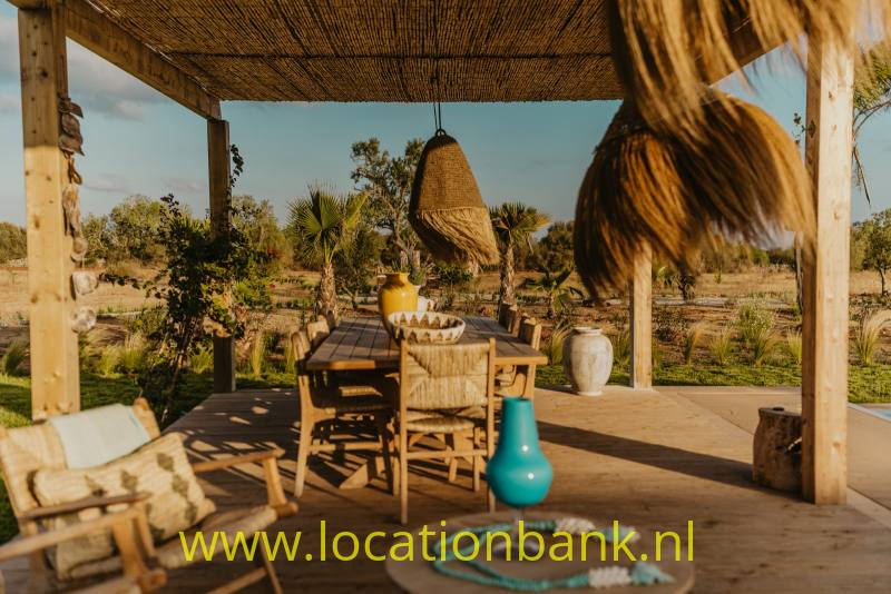 Overdekt houten terras met een rieten dak, eettafel, turquoise vaas en droge tuin op de achtergrond.