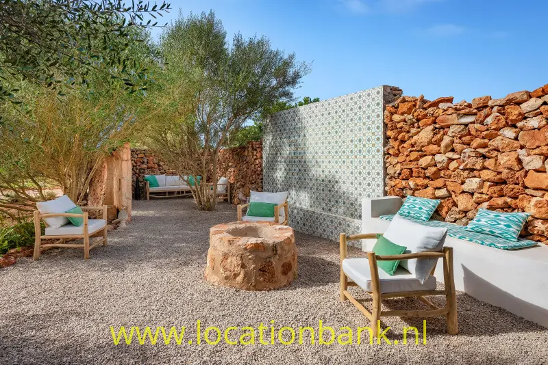 Grindbinnenplaats met een betegelde wand, natuurstenen muren, olijfbomen, houten fauteuils en een ronde stenen tafel.