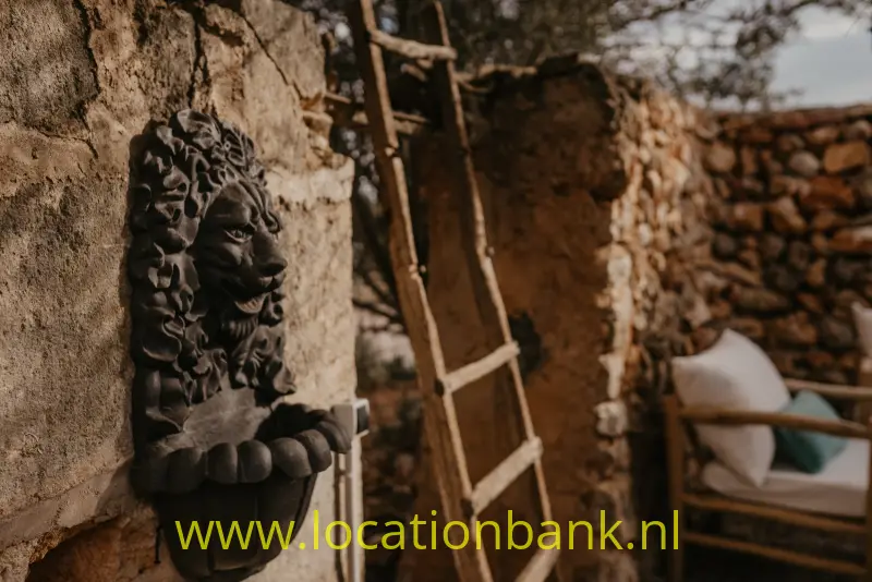 Een donker leeuwenkopreliëf hangt aan een verweerde stenen muur naast een houten ladder.