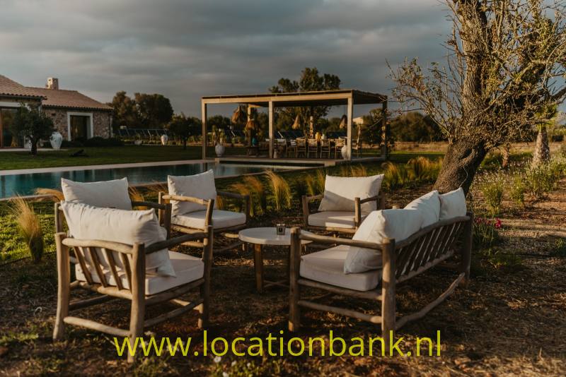 Houten tuinmeubelen met witte kussens staan naast een zwembad, met een overdekte pergola op de achtergrond.