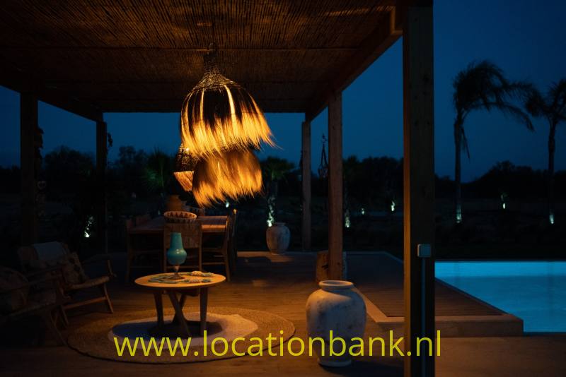 Een gevlochten hanglamp verlicht een rond tafeltje op een overdekt terras naast een zwembad.