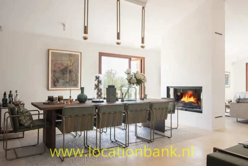Lange eettafel met meerdere stoelen, drie hanglampen, een schilderij en een brandende haard.
