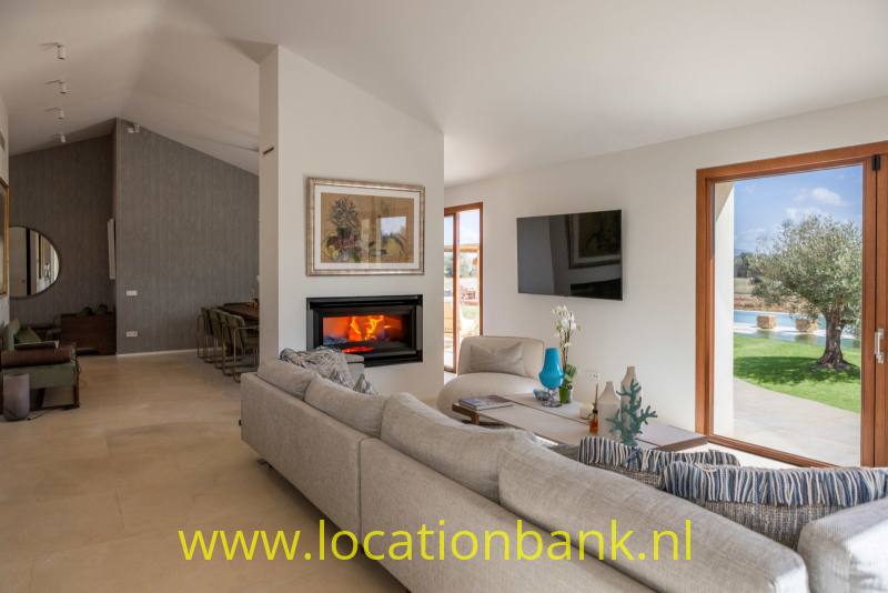 Woonkamer met grote grijze hoekbank, open haard, televisie en glazen deuren naar een tuin met een olijfboom.