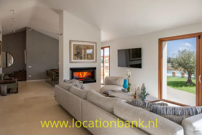 Woonkamer met grote grijze hoekbank, open haard, televisie en glazen deuren naar een tuin met een olijfboom.