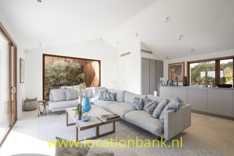Ruime woonkamer met een grijze hoekbank, lage salontafel en grote ramen naast een open keuken.