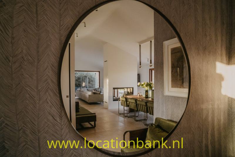 Grote ovale spiegel toont een lichte woonkamer met een eettafel, barkrukken en hanglampen.