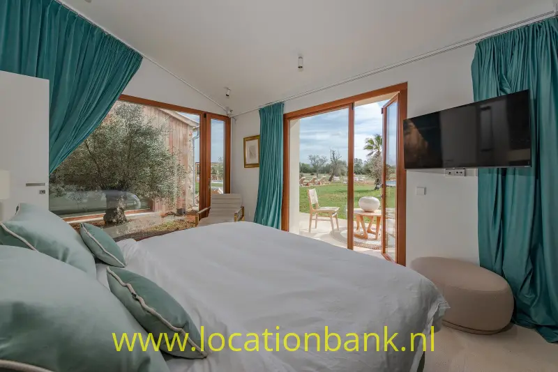 Slaapkamer met opgemaakt bed, turquoise gordijnen en openslaande glazen deuren naar een groene tuin.