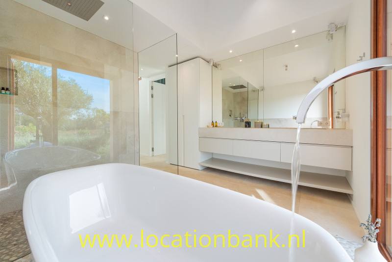 Vrijstaand wit bad op de voorgrond van een moderne badkamer met dubbele wastafel en glazen douchewand.