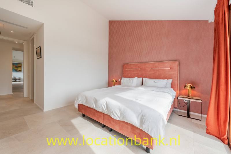 Een tweepersoonsbed met wit beddengoed staat voor een roodroze wand, naast een open doorgang.