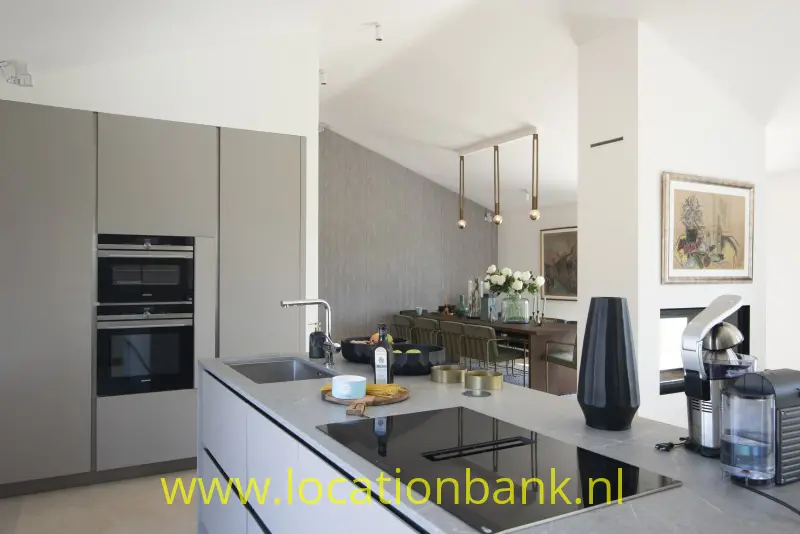 Kookeiland met donker werkblad, ingebouwde ovens, hanglampen en een aangrenzende eethoek.