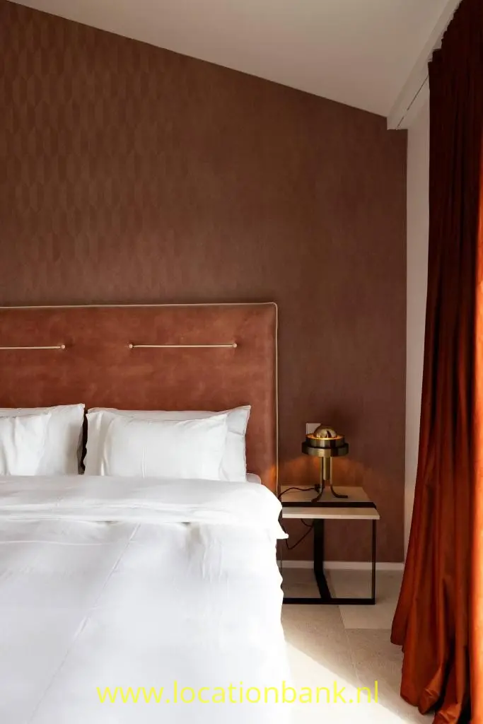Bed met wit beddengoed tegen een bruine wand, naast een nachtkastje en lange rode gordijnen.