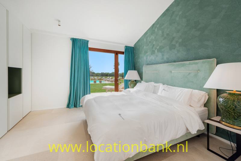 Slaapkamer met een opgemaakt tweepersoonsbed, een groen getextureerde wand en turquoise gordijnen naast het raam.