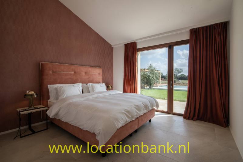 Slaapkamer met een opgemaakt tweepersoonsbed, lange gordijnen en glazen deuren naar een groene buitenruimte.