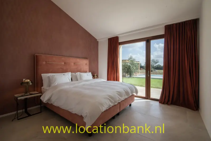 Slaapkamer met een opgemaakt tweepersoonsbed, lange gordijnen en glazen deuren naar een groene buitenruimte.