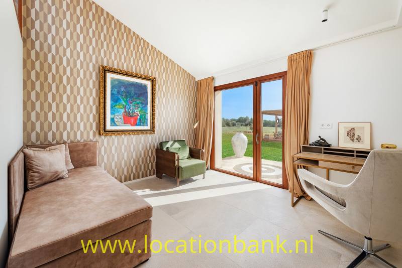 Woonkamer met een hoekbank, geometrisch behang, een bureau en openslaande glazen deuren naar de tuin.