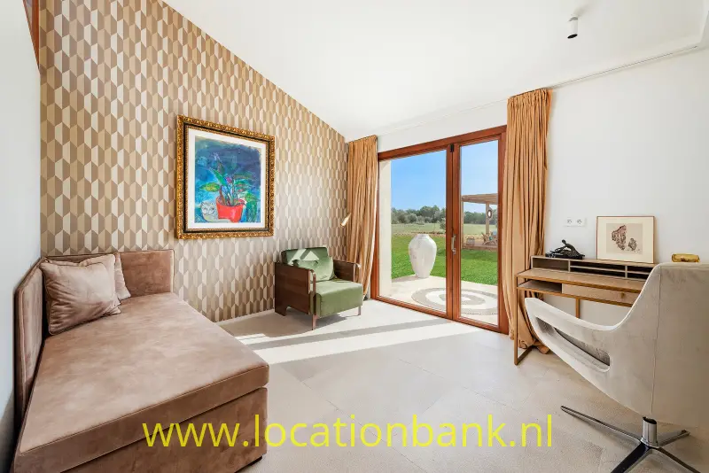 Woonkamer met een hoekbank, geometrisch behang, een bureau en openslaande glazen deuren naar de tuin.