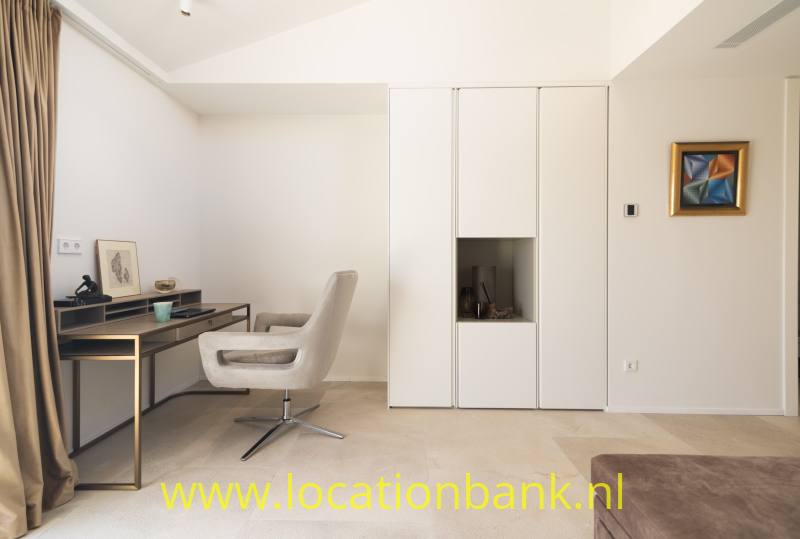 Witte kamer met een bureau, gestoffeerde draaistoel, hoge ingebouwde kasten en ingelijst kunstwerk.