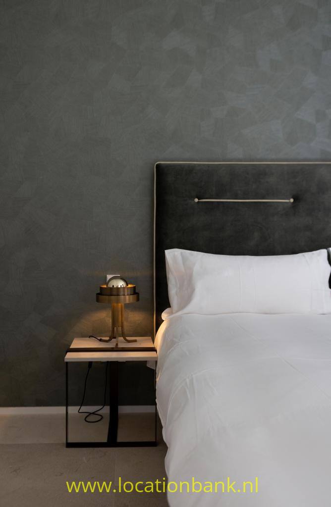 Een bed met wit beddengoed staat naast een zwart nachtkastje met tafellamp tegen een donkere wand.
