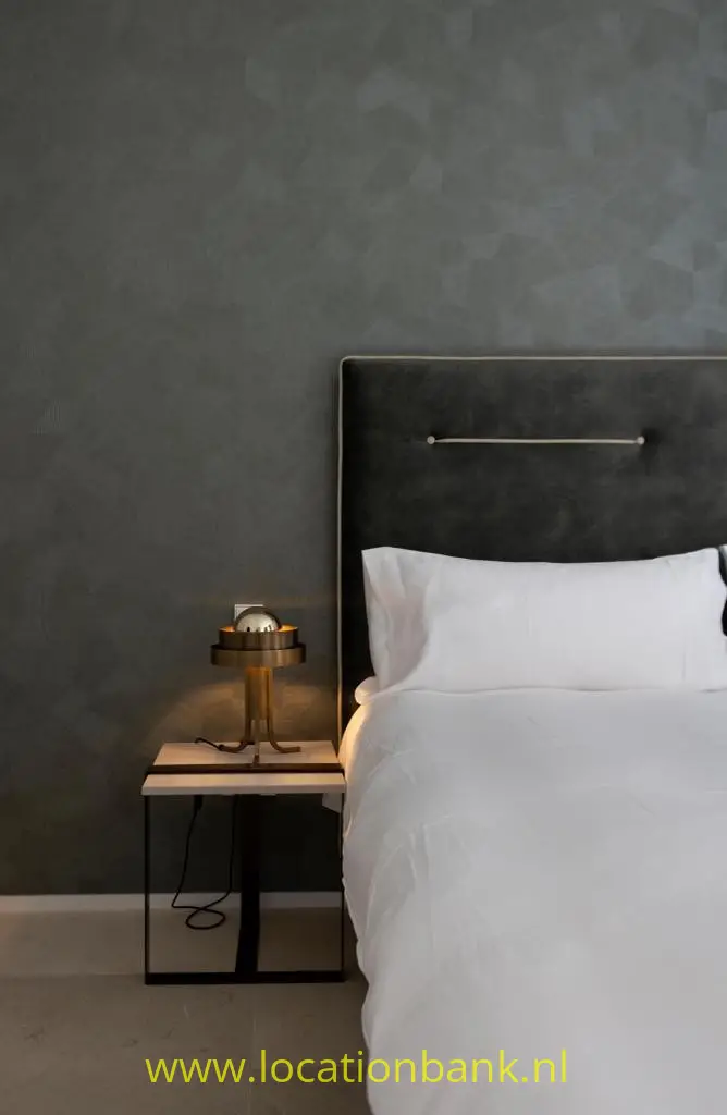 Een bed met wit beddengoed staat naast een zwart nachtkastje met tafellamp tegen een donkere wand.
