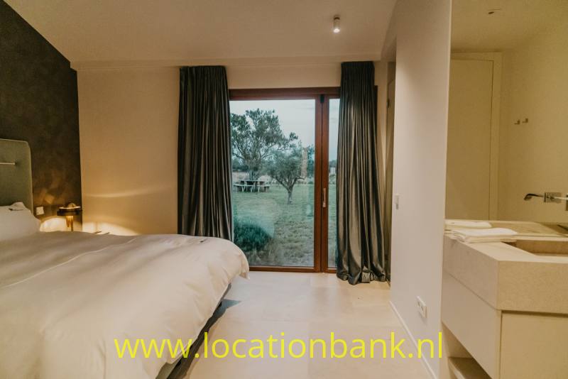 Slaapkamer met een opgemaakt bed, donkere gordijnen en een glazen deur met uitzicht op bomen en gras.