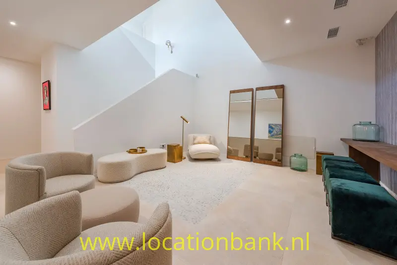 Beigekleurige fauteuils staan rond een lage, ovale poef, naast een witte trap en twee hoge spiegels.