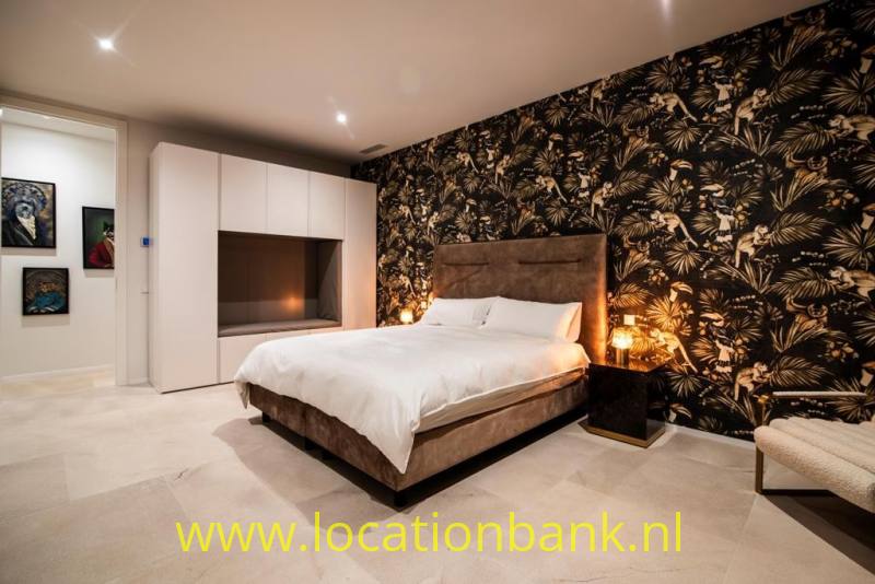 Slaapkamer met een groot bed, donker botanisch behang, witte kasten en warme verlichting.
