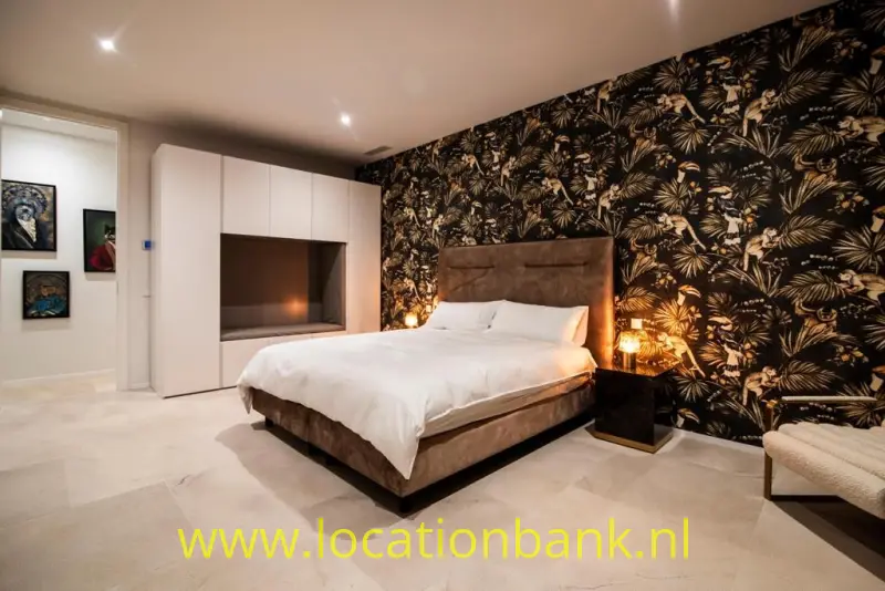 Slaapkamer met een groot bed, donker botanisch behang, witte kasten en warme verlichting.