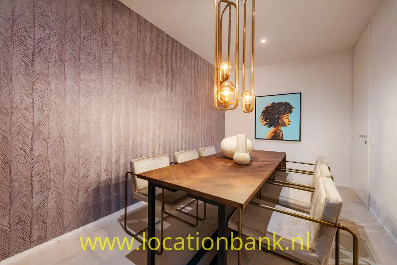 Houten eettafel met stoelen onder glazen hanglampen, naast een wand met patroonbehang en een portret.