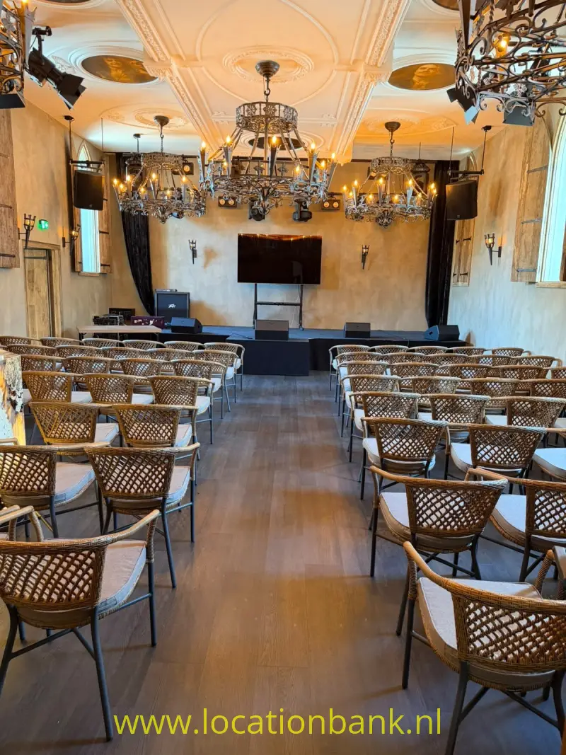 Rijen gevlochten stoelen staan gericht op een klein podium onder meerdere kroonluchters en sierlijke plafonddecoraties.