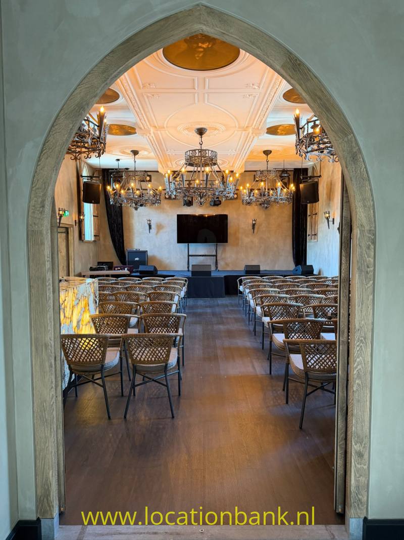 Een boogvormige doorgang leidt naar een zaal met rijen stoelen, kroonluchters en een gedecoreerd plafond.