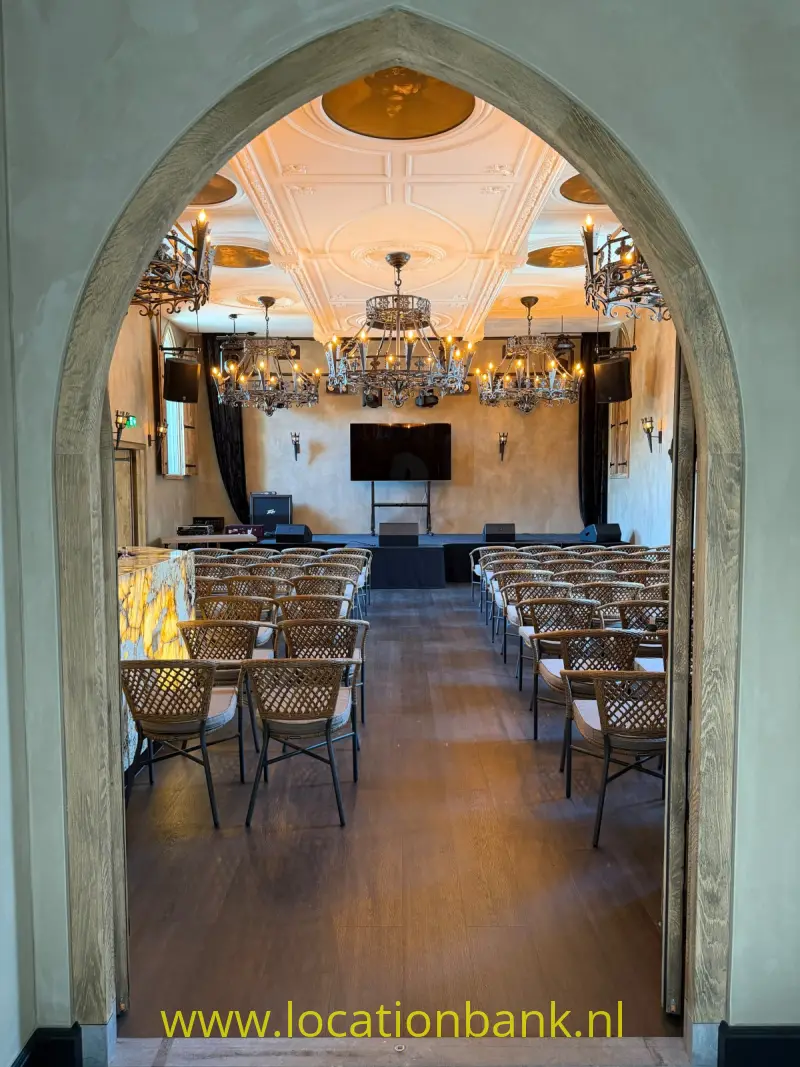 Een boogvormige doorgang leidt naar een zaal met rijen stoelen, kroonluchters en een gedecoreerd plafond.