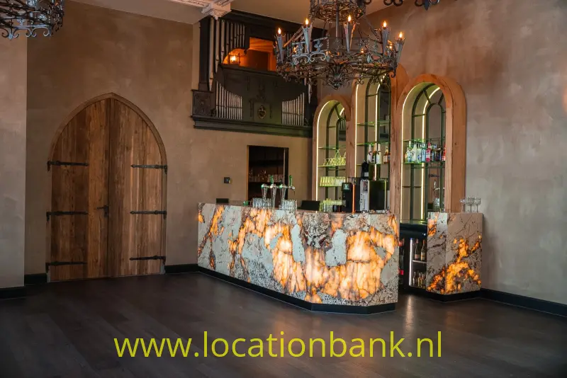 Bar met een verlichte stenen toonbank, hoge boogramen, kroonluchters en een grote houten boogdeur.