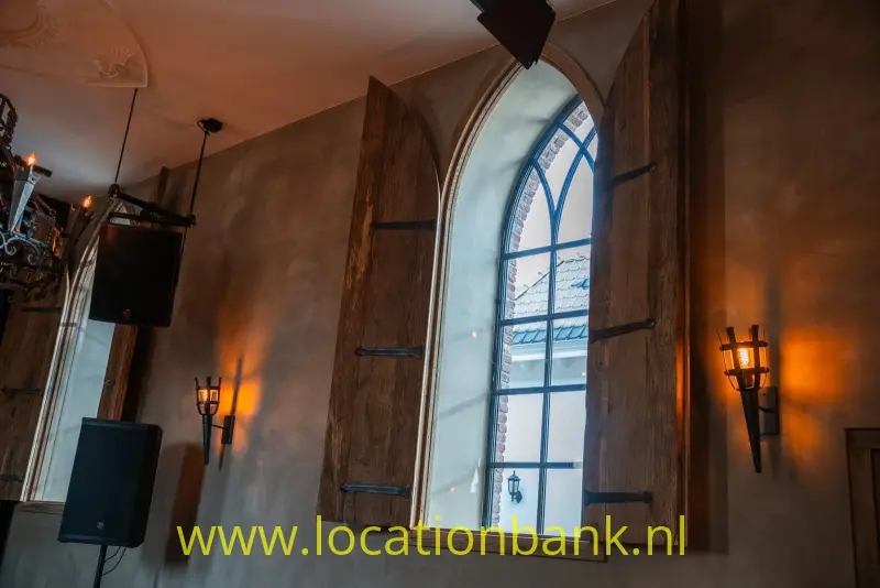 Een hoog boograam met houten luiken, wandlampen en een kroonluchter in een interieur.