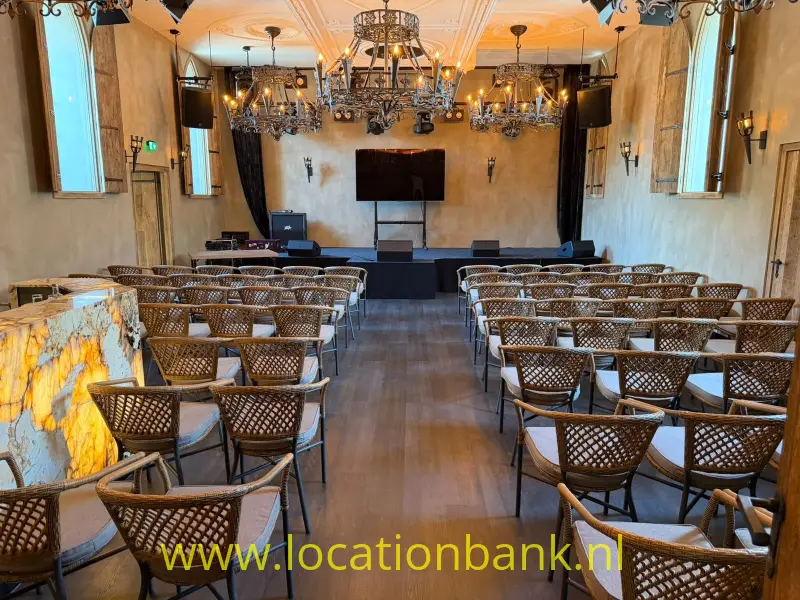 Zaal met rijen gevlochten stoelen, kroonluchters, een podium en hoge ramen langs de wanden.