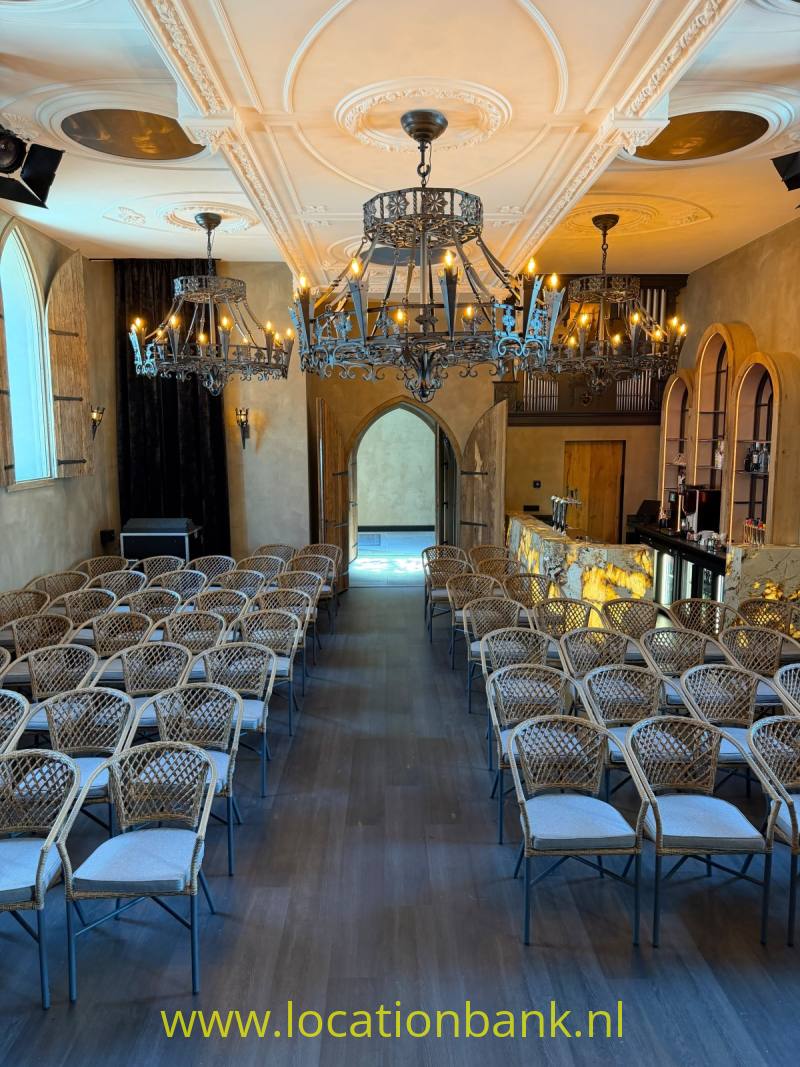 Zaal met rijen gevlochten stoelen, meerdere kroonluchters, boogvormige doorgangen en een verlichte bar achterin.