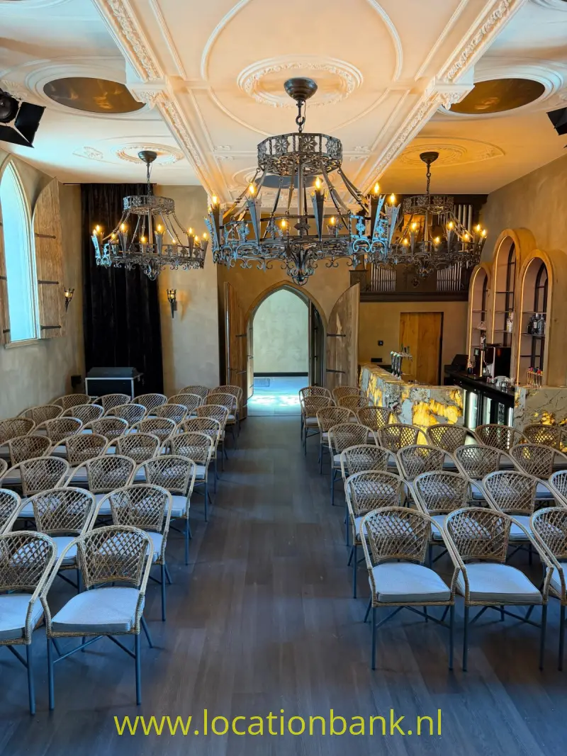 Zaal met rijen gevlochten stoelen, meerdere kroonluchters, boogvormige doorgangen en een verlichte bar achterin.