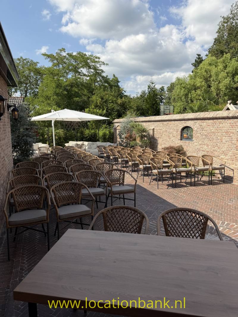 Buitenterras met gevlochten stoelen en tafels, een witte parasol, een bakstenen muur en bomen.