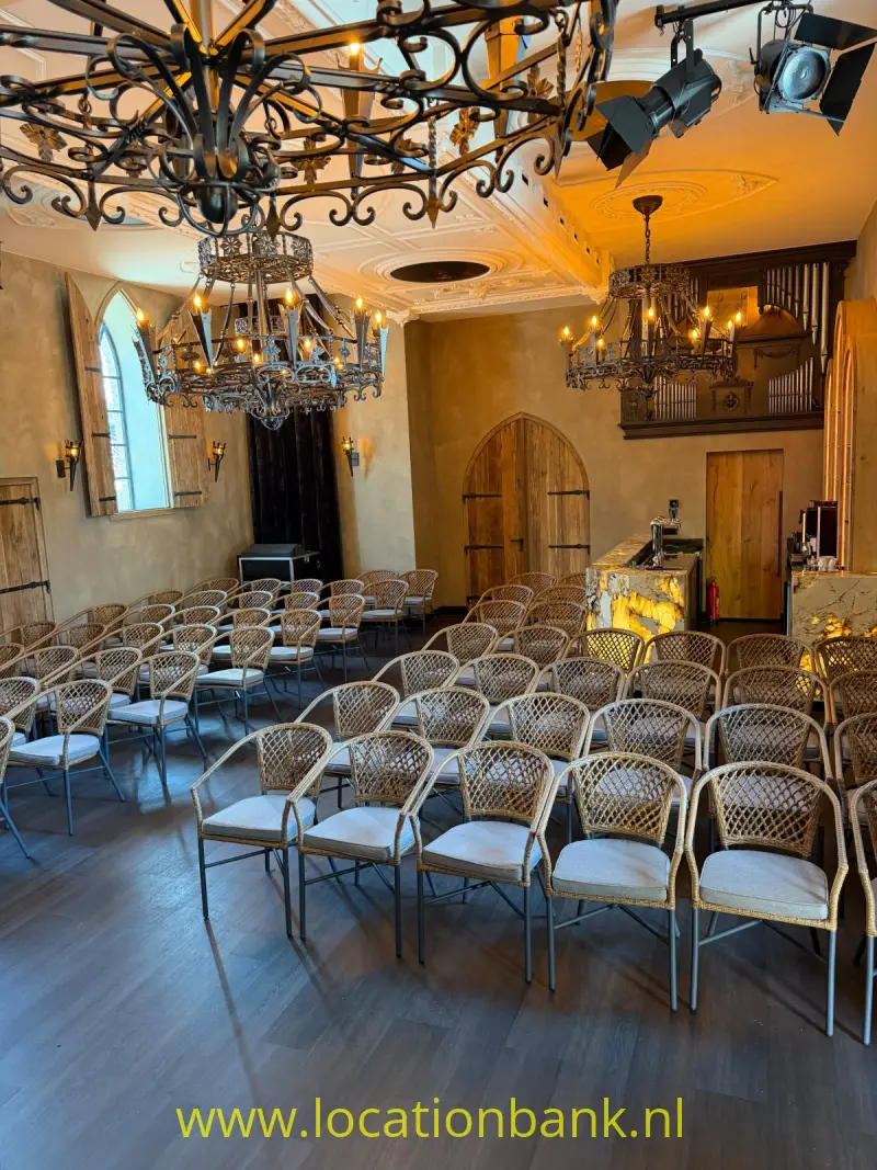 Rijen gevlochten stoelen staan in een zaal met kroonluchters, boogramen en een versierd plafond.