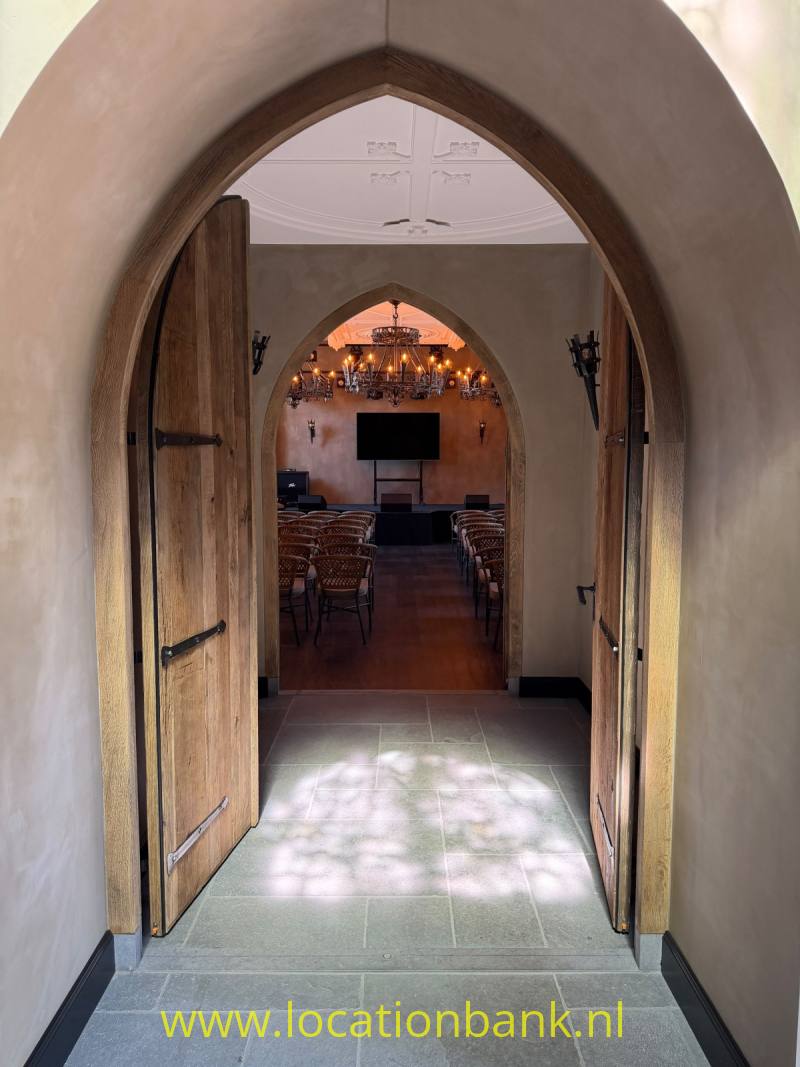Een openstaande houten boogdeur biedt zicht op een zaal met tafels, stoelen, kroonluchters en een scherm.