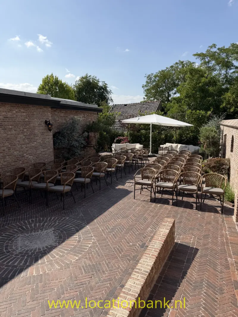Bakstenen terras met rijen gevlochten stoelen, tafels en witte parasols, omgeven door lage bakstenen muren en bomen.