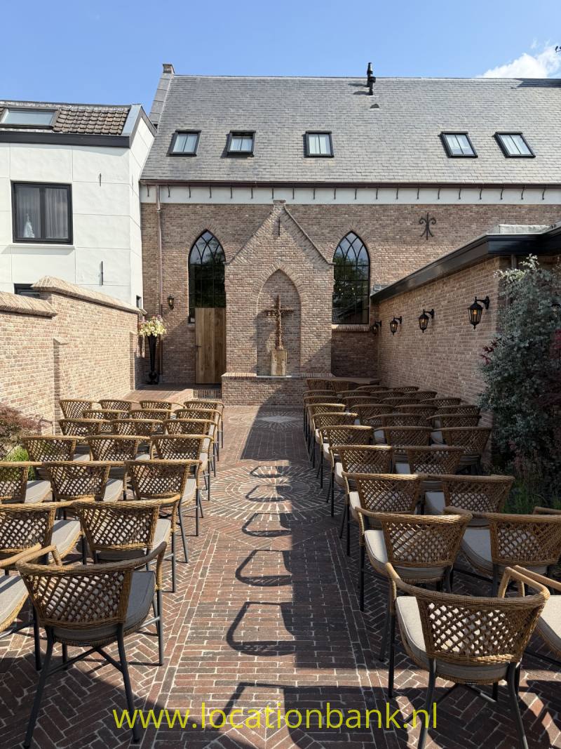 Bakstenen gebouw met hoge spitsboogvensters en een terras met rijen gevlochten stoelen.