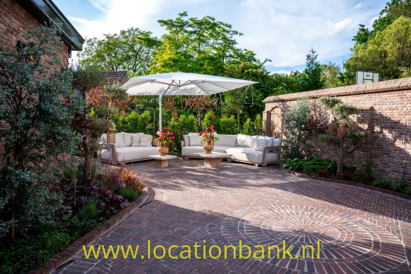 Een bakstenen binnenplaats heeft een grote parasol, witte zitbanken, bloeiende planten en omringende bakstenen muren.