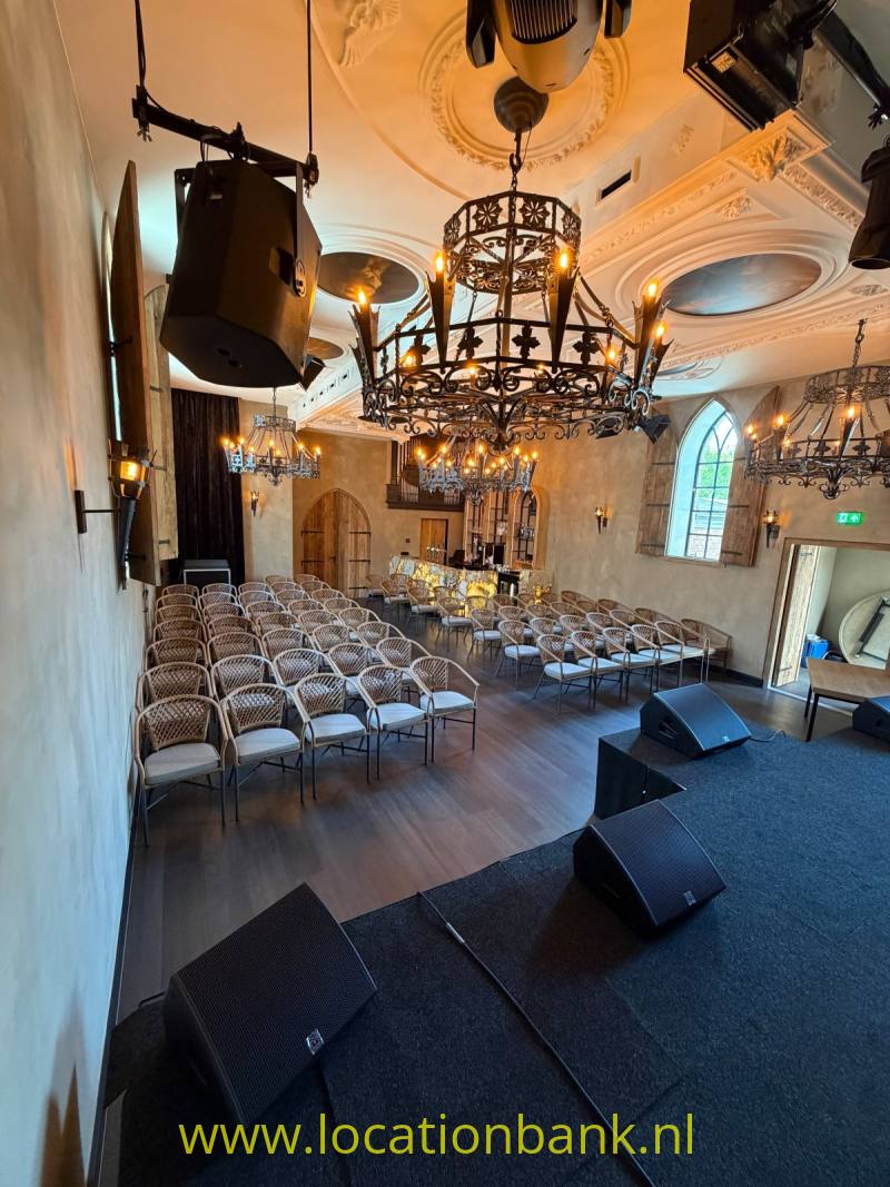 Meerdere sierlijke kroonluchters hangen boven rijen stoelen en een podium met luidsprekers in een gedecoreerde zaal.