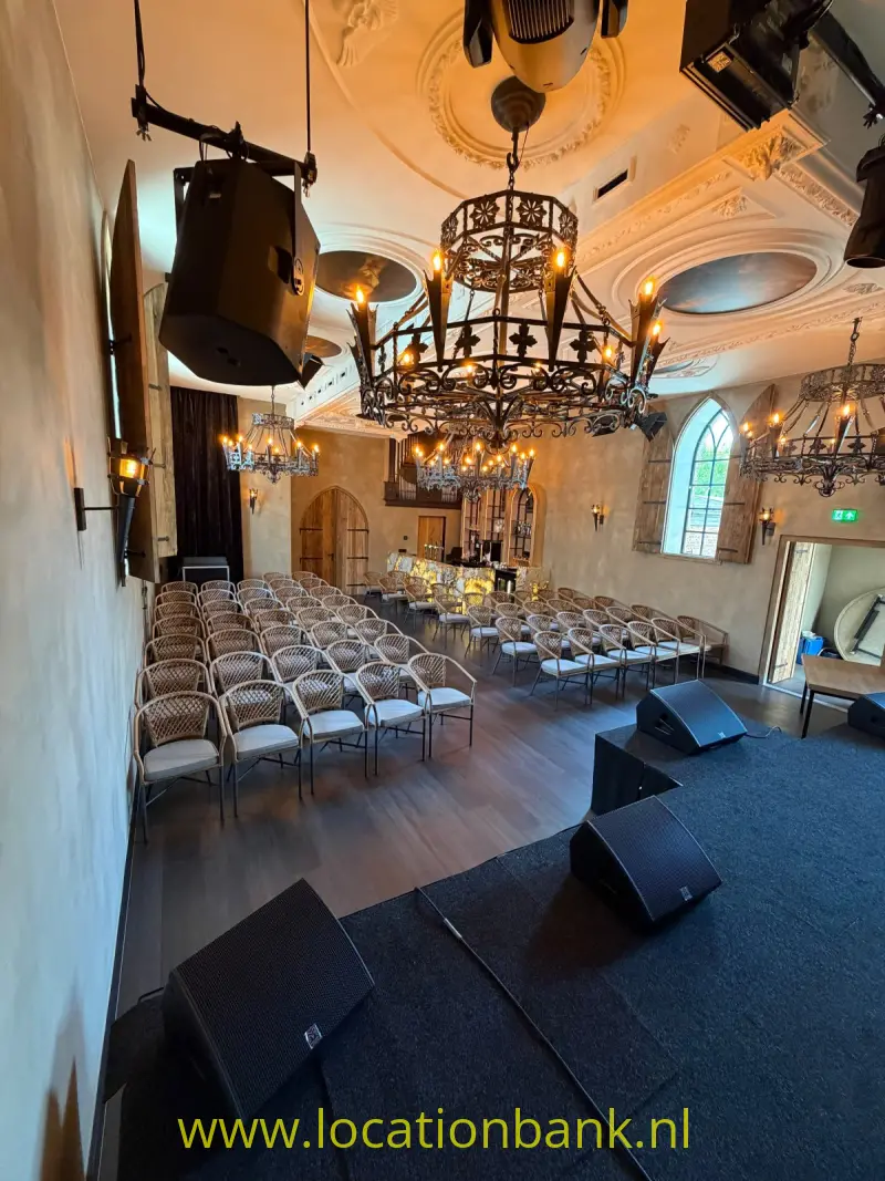 Meerdere sierlijke kroonluchters hangen boven rijen stoelen en een podium met luidsprekers in een gedecoreerde zaal.