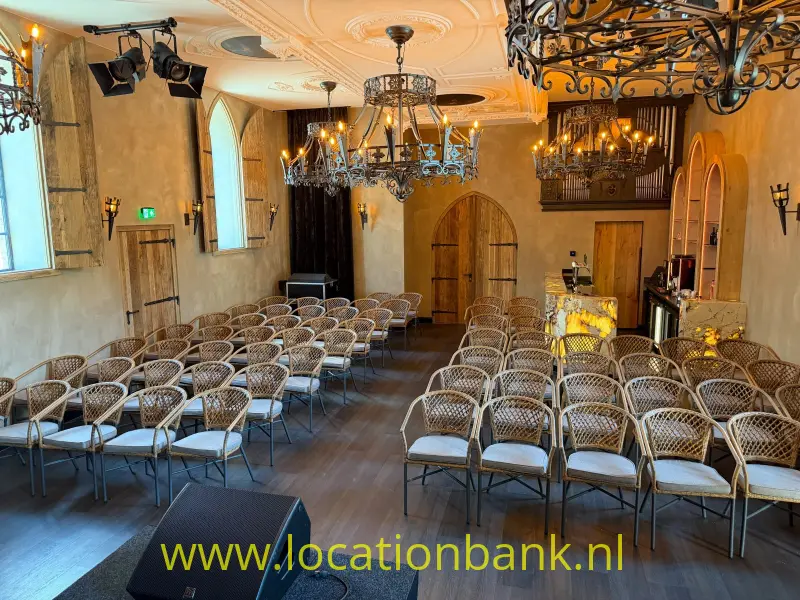 Rijen gevlochten stoelen staan in een zaal met kroonluchters, sierlijke plafonddecoratie en houten deuren.