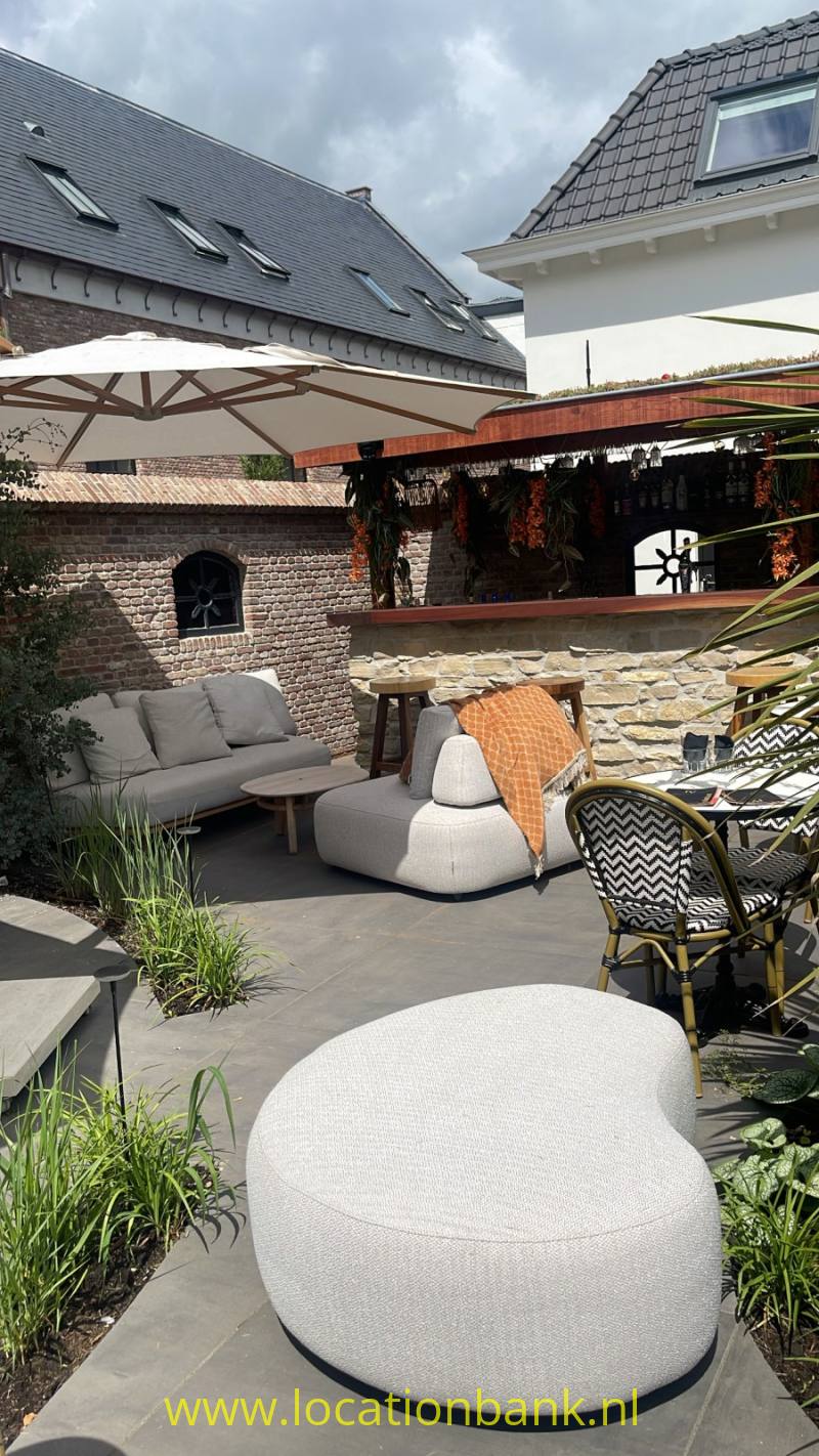 Een grote ronde, lichtgekleurde zitpoef staat op een terras tussen tuinmeubilair en parasols.