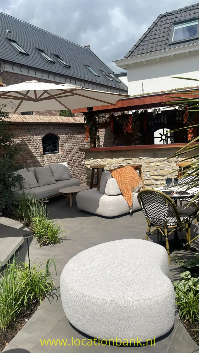 Een grote ronde, lichtgekleurde zitpoef staat op een terras tussen tuinmeubilair en parasols.